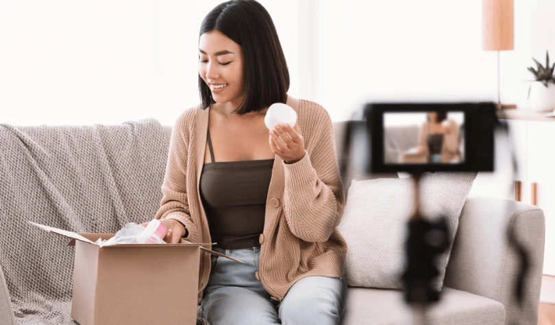 A smiling woman recording content on a couch with a camera and delivery boxes.