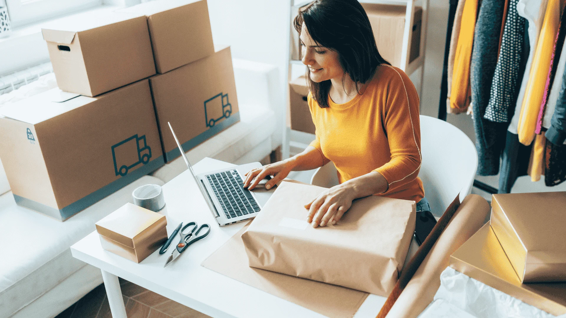 A woman in a yellow shirt prepares packages for delivery while working at a laptop.
